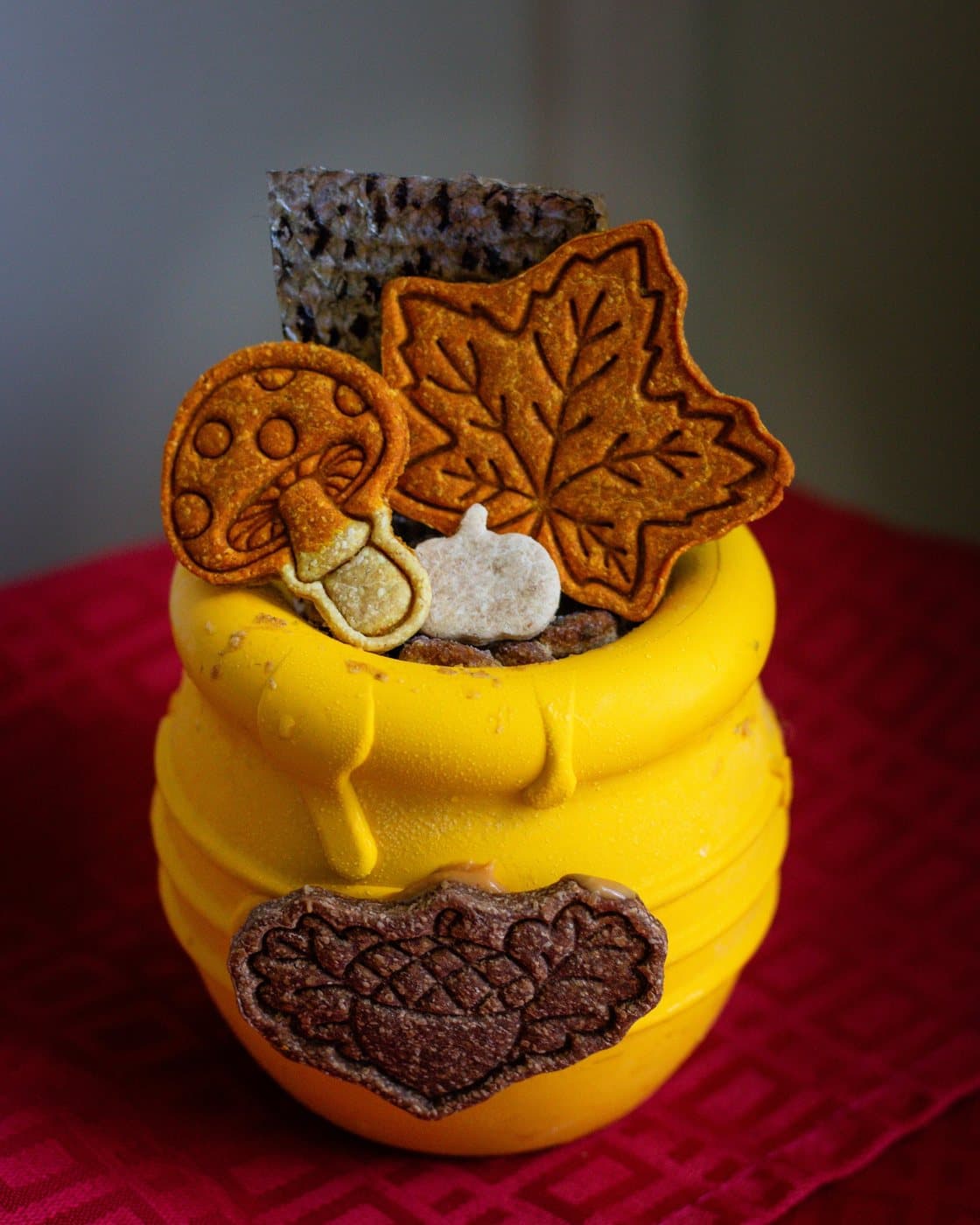 Biscuits pour chiens d'automne : une feuille d'érable dorée, un champignon, une citrouille et un gland garnissent un pot de miel jaune posé sur une nappe rouge